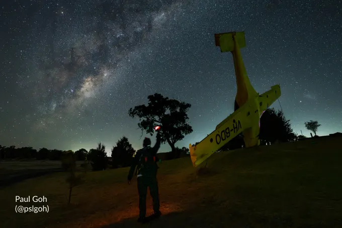 A photographer under the Milky Way beside a yellow aircraft, holding up a red light