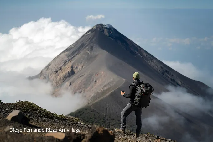 A hiker with a camera backpack looking at a volcano above the clouds